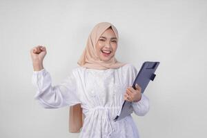 Excited young Asian Muslim woman wearing white dress and hijab holding a document, cheering and celebrating. Isolated by white background. Ramadan and Eid Mubarak concept photo