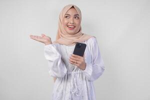 Smiling Asian woman wearing hijab and white dress using smartphone while pointing to the copy space on her right side, standing over isolated white background. Ramadan and Eid Mubarak concept photo