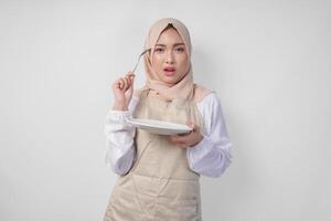 Confused young Asian Muslim woman in hijab and cream apron holding spoon and empty plate with copy space over it, thinking hard what food menu to eat for iftar. Ramadan concept photo