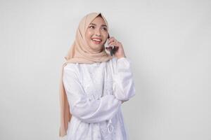 Happy Asian Muslim woman wearing white dress and hijab holding her smartphone and making a call to family or friend, isolated by white background. Ramadhan and Eid Mubarak concept photo