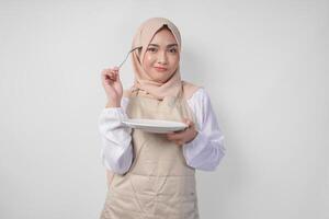 Confused young Asian Muslim woman in hijab and cream apron holding spoon and empty plate with copy space over it, thinking hard what food menu to eat for iftar. Ramadan concept photo