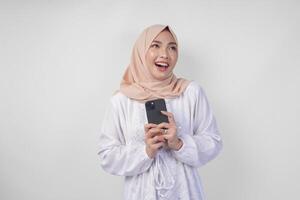 Thoughtful young Asian Muslim woman wearing white dress and hijab holding phone while thinking and finding idea with happy expression, isolated on white background. Ramadan and Eid Mubarak concept photo