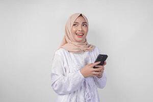 Thoughtful young Asian Muslim woman wearing white dress and hijab holding phone while thinking and finding idea with happy expression, isolated on white background. Ramadan and Eid Mubarak concept photo