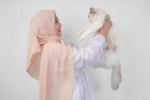 retrato de un hermosa joven musulmán mujer vistiendo blanco vestir y hiyab, sonriente y participación un blanco muñeca de trapo gato terminado aislado blanco antecedentes foto