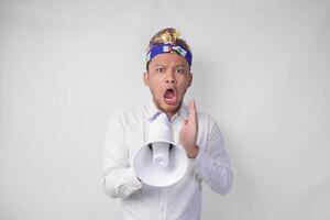 Overjoyed Balinese man in white shirt and traditional headdress shouting at megaphone feeling excited, isolated by white background photo