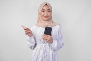 Smiling Asian woman wearing hijab and white dress using smartphone while pointing to the copy space on her right side, standing over isolated white background. Ramadan and Eid Mubarak concept photo