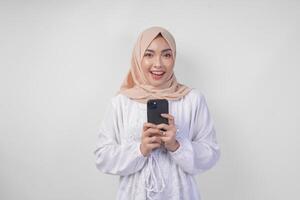 emocionado asiático musulmán mujer vistiendo blanco vestir y hijab participación su teléfono inteligente mientras sonriente alegremente, aislado por blanco antecedentes. ramadhan y eid Mubarak concepto foto