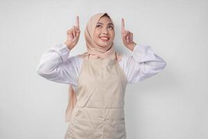 Overjoyed Asian woman wearing hijab and cream apron pointing up to the copy space above, standing over isolated white background photo