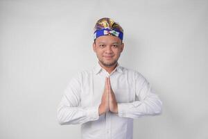 Smiling young Balinese man wearing traditional headdress called udeng doing greeting or welcome gesture, isolated over white background photo