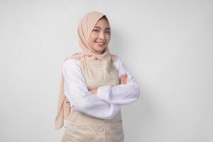 Young Asian Muslim woman wearing hijab and cream apron smiling to the camera with crossed arms pose full of confidence photo