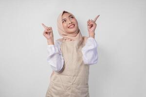 Overjoyed Asian woman wearing hijab and cream apron pointing up to the copy space above, standing over isolated white background photo