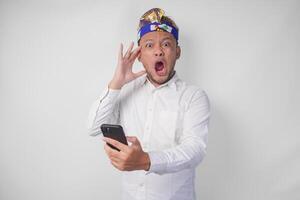 Angry Balinese man in white shirt and traditional headdress feeling mad and upset while using smartphone photo