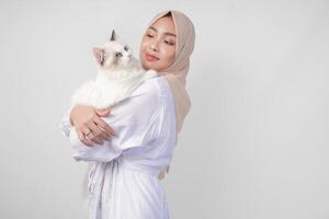 retrato de un hermosa joven musulmán mujer vistiendo blanco vestir y hiyab, sonriente y participación un blanco muñeca de trapo gato terminado aislado blanco antecedentes foto