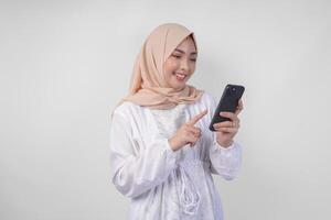 Excited Asian Muslim woman wearing white dress and hijab holding her smartphone while smiling cheerfully, isolated by white background. Ramadhan and Eid Mubarak concept photo