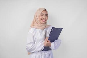 Smiling young Asian Muslim woman wearing white dress and hijab holding a document, Isolated by white background. Ramadan and Eid Mubarak concept photo