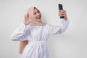 Portrait of Asian Muslim woman wearing white dress and hijab holding her smartphone to do a call to family or friend, isolated by white background. Ramadhan and Eid Mubarak concept photo