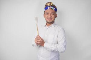 Balinese man wearing traditional headdress called udeng doing paying respect gesture while holding praying incense on isolated white background photo