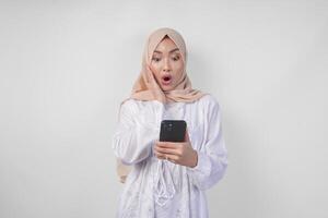 Young Asian Muslim woman wearing white dress and hijab showing shocked expression on her face while holding smartphone, surprised after reading news or gossip, isolated by white background photo