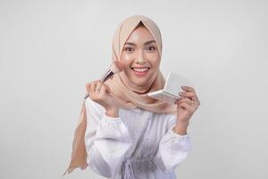 Beautiful young Asian Muslim woman wearing white dress and hijab applying make up using brush. Fashion and cosmetics beauty concept photo