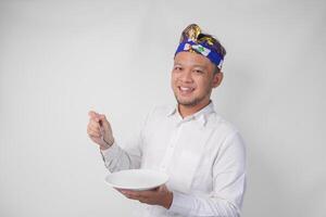 retrato de un joven balinés hombre en blanco camisa y tradicional tocado demostración y presentación un vacío plato con Copiar espacio mientras sonriente alegremente a el cámara foto