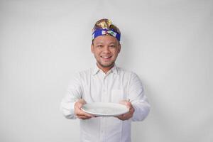 Smiling young Balinese man in white shirt and traditional headdress showing and presenting an empty plate with copy space photo