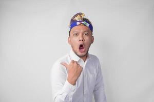 Surprised Balinese man in white shirt and traditional headdress called udeng pointing to the copy space on the left and right side photo