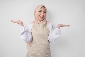 Excited young Asian Muslim woman in hijab and cream apron pointing to the empty copy space on the left and right side. Ramadan advertisement concept photo