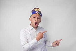 Surprised Balinese man in white shirt and traditional headdress called udeng pointing to the copy space on the left and right side photo