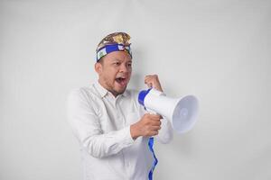 Overjoyed Balinese man in white shirt and traditional headdress shouting at megaphone feeling excited, isolated by white background photo