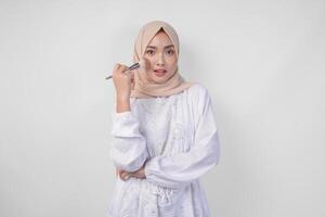 hermosa joven asiático musulmán mujer vistiendo blanco vestir y hijab aplicando hacer arriba utilizando cepillar. Moda y productos cosméticos belleza concepto foto