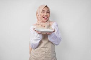 hermosa joven asiático musulmán mujer en hijab y crema delantal presentación un vacío plato con Copiar espacio terminado él, demostración el comida menú para iftar foto