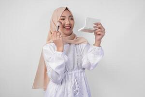 Beautiful young Asian Muslim woman wearing white dress and hijab applying make up using brush. Fashion and cosmetics beauty concept photo