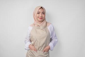 Smiling young Asian Muslim woman in a veil hijab and cream apron doing a formal pose with hands tucked in pockets photo