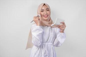 Beautiful young Asian Muslim woman wearing white dress and hijab applying make up using brush. Fashion and cosmetics beauty concept photo