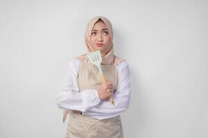 Thoughtful young Asian Muslim woman in hijab and cream apron holding spatula and kitchen cooking utensils, thinking hard what food menu to cook photo