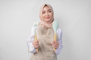 emocionado joven asiático musulmán mujer en un velo hijab y crema delantal sonriente a el cámara mientras participación espátula y cocina Cocinando utensilios foto