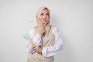 Thoughtful young Asian Muslim woman in hijab and cream apron holding spatula and kitchen cooking utensils, thinking hard what food menu to cook photo