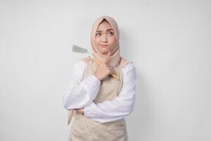 Thoughtful young Asian Muslim woman in hijab and cream apron holding spatula and kitchen cooking utensils, thinking hard what food menu to cook photo