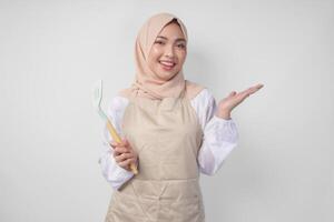 Happy young Asian Muslim woman in a veil hijab and cream apron holding spatula while pointing to the copy space beside her photo