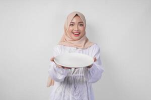 Beautiful young Asian Muslim woman in hijab presenting an empty plate with copy space over it, showing the food menu for iftar photo
