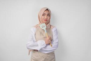 Thoughtful young Asian Muslim woman in hijab and cream apron holding spatula and kitchen cooking utensils, thinking hard what food menu to cook photo