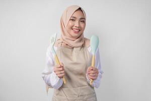 emocionado joven asiático musulmán mujer en un velo hijab y crema delantal sonriente a el cámara mientras participación espátula y cocina Cocinando utensilios foto