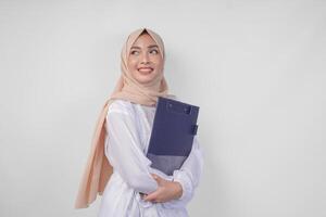 sonriente joven asiático musulmán mujer vistiendo blanco vestir y hijab participación un documento, aislado por blanco antecedentes. Ramadán y eid Mubarak concepto foto