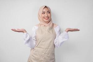 Excited young Asian Muslim woman in hijab and cream apron pointing to the empty copy space on the left and right side. Ramadan advertisement concept photo