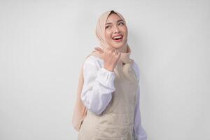 Excited young Asian Muslim woman in hijab and cream apron pointing to the empty copy space on the left and right side. Ramadan advertisement concept photo