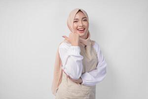 Excited young Asian Muslim woman in hijab and cream apron pointing to the empty copy space on the left and right side. Ramadan advertisement concept photo