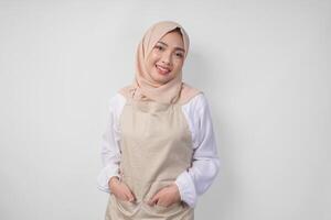 Smiling young Asian Muslim woman in a veil hijab and cream apron doing a formal pose with hands tucked in pockets photo