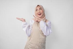 Excited young Asian Muslim woman in hijab and cream apron pointing to the empty copy space on the left and right side. Ramadan advertisement concept photo