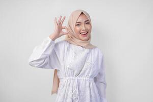 Portrait of Asian Muslim woman in white dress and hijab making an okay, good, nice sign gesture using fingers. Ramadan and Eid Mubarak concept photo