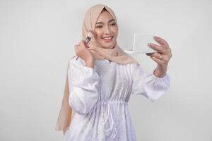 Beautiful young Asian Muslim woman wearing white dress and hijab applying make up using brush. Fashion and cosmetics beauty concept photo